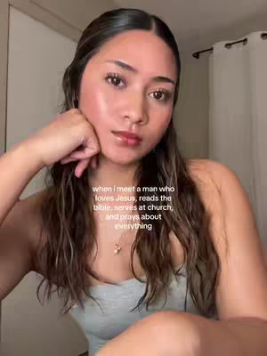 The image shows a young woman in a close-up shot. She has long, dark hair and is wearing a gray tank top. The background is a plain, light-colored wall. Text overlay at the bottom.