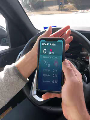 A person is holding a smartphone that is displaying a heart rate monitor app while driving. The person's hands, the steering wheel, and a glimpse of the car's interior are visible. Outside, a cityscape is slightly visible through the windshield.