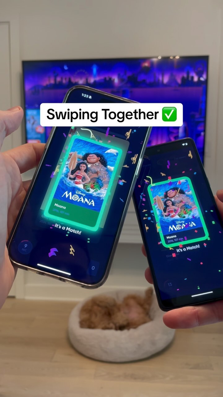 A person holds two smartphones with dating app interfaces showing a movie title and a green checkmark. In the background, a blurred television is visible. A fluffy dog bed sits on the floor nearby.