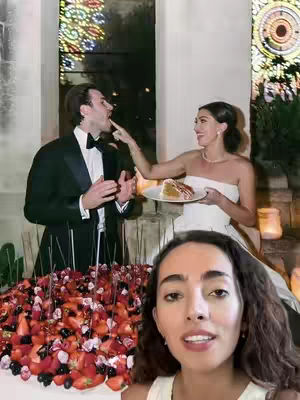 A woman in the foreground looks at the camera with a smile. Behind her, a wedding scene unfolds: a couple interacts with a cake, with a unique arrangement of strawberries.