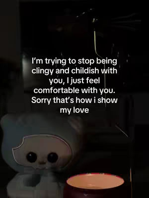 A dimly lit scene, featuring a light blue plush toy resembling a character with large eyes in the foreground, positioned slightly left of center. In front of the toy, a small, lit candle with a red container glows, providing the primary light source. Above the toy and slightly behind, text is overlaid in white, conveying a message. The background is dark, with vague shapes.