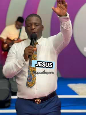 A man in a white dress shirt and blue pants is the central figure in the image, raising his right hand in the air. The image is set in what appears to be a church, with another person visible playing a guitar in the background. Overlayed on the image is a text box that says 'JESUS'.