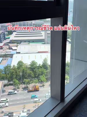 The image shows a view from a high-rise window, looking out over a city street. Visible is a multilane highway with moving vehicles. Buildings and trees are visible in the background. Overlaid on the image is Thai text in red. The window frame is to the right with the sky and background in view on the left.