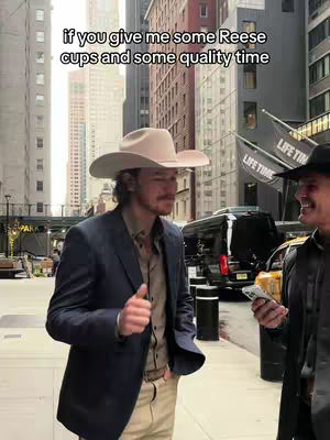 Two men in a city setting, likely New York. One, wearing a cowboy hat and a suit jacket, is the focus, next to him is another male wearing a black leather jacket, and the background includes tall buildings, and the Life TIme Logo.