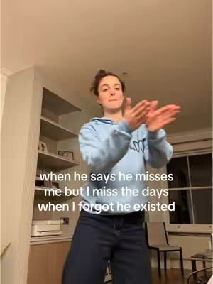 A young woman is standing in a well-lit room with her arms raised in front of her, palms facing each other as if clapping. She is wearing a light blue hoodie and dark-colored pants. The background suggests a domestic setting.