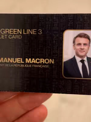 A photograph of a black transit card-like design held in a hand, featuring details in gold and a photo of a man in a suit. The top section of the card has gold text that reads 'GREEN LINE 3' and below that, 'TICKET CARD'. The lower section of the card features the text 'EMMANUEL MACRON' and below that, the text 'PRESIDENT DE LA RÉPUBLIQUE FRANÇAISE' and a small portrait of a man with short brown hair in a business suit and black tie. The background of the card is a dark, possibly textured, material, while the hand holding the card is slightly visible in the foreground.