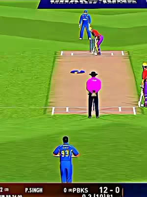 An overhead view of a cricket game in progress. The field is green, with a brown pitch at the center. Players in various colored uniforms are positioned around the pitch. A large scoreboard is visible at the bottom, displaying game stats.