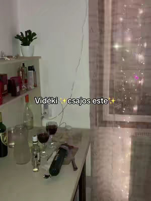 An indoor scene shows a countertop with items like wine glasses, bottles, and a microphone. The background includes shelves with toiletries and a window with blurred lights.