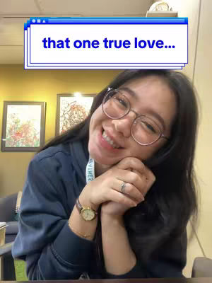 A young woman with long dark hair and glasses smiles warmly at the camera. She's wearing a dark blue hoodie. Above her head is a speech bubble displaying 'that one true love...'. The background includes a soft, yellow-toned wall and artwork.