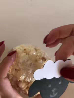 The image features two hands with red nail polish holding a circular object wrapped in clear plastic. A cutout of a black sheep with a white cloud shape is on the right. The background is a solid, light-colored surface.