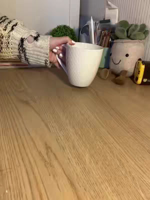 A hand wearing a knitted sweater holds a white mug on what looks like a wooden desk, with a glimpse of study-related items in the background.
