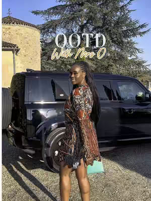 A woman stands in front of a black SUV, smiling at the camera. She wears a patterned dress and has long, dark hair. The background includes a building and foliage. The text overlay reads 'OOTD With Mrs. O'.