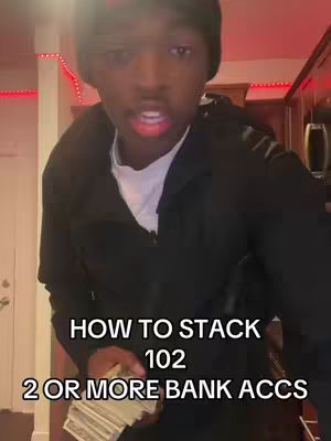 A young Black man in a black hoodie and beanie speaks directly to the camera. Text overlays read: "HOW TO STACK 102 2 OR MORE BANK ACCS." He holds a wad of cash.
