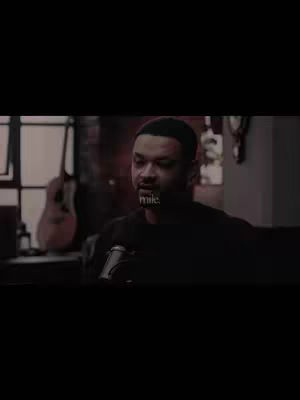 A man is seated in front of a microphone, looking to his left. The lighting is dim with some natural light coming from a window to the left; a guitar is slightly visible in the background. Text overlayed on the image reads, "mike."