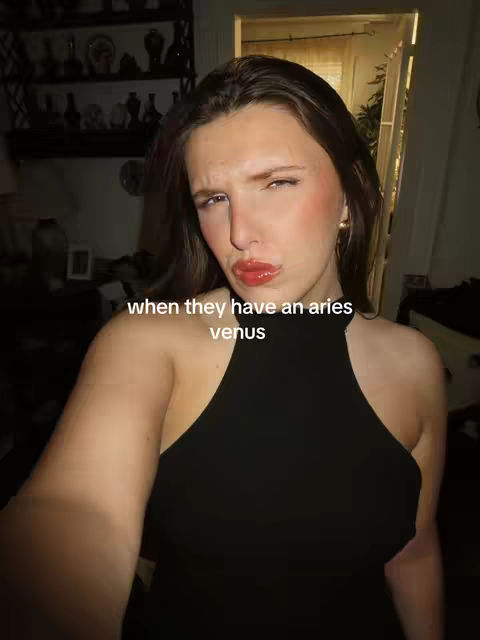 A selfie-style image features a young woman with long dark hair, looking directly at the camera. She's indoors, with a dark top and vibrant makeup. The background has a shelf with decorations, and some light filtered through a window or doorway. Text overlayed on the image reads, 'when they have an aries venus'.