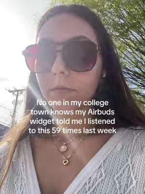 A selfie-style shot of a young woman outdoors. She is wearing large sunglasses and various necklaces, with text overlayed at the bottom of the image. The background suggests an urban or suburban setting with greenery.
