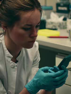 A woman in a white lab coat and teal gloves looks down at a phone she is holding. The background is slightly blurry but shows a lab setting.