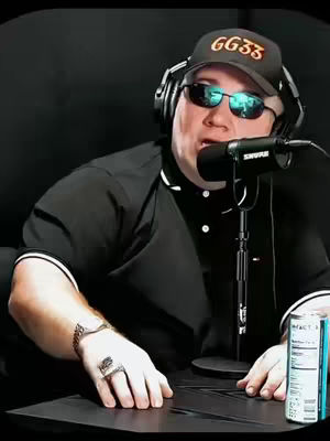A man wearing a black polo shirt, cap and sunglasses is sitting behind a desk with a microphone and can. The background is black. The man is holding the microphone close to his mouth.