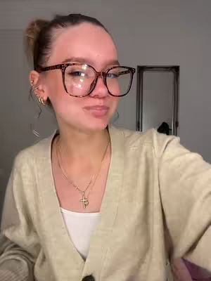 A woman with fair skin, wearing large-framed glasses and a light-colored cardigan, looks towards the camera. She has light brown hair pulled back in a bun and is wearing a gold necklace with a cross pendant. The background is a neutral, blurry setting with a mirror visible on the right.