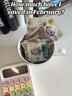 The image shows a top-down view of a white mug overflowing with a collection of British banknotes. A white calculator with colorful buttons sits to the left of the mug, which is positioned against a white surface. Along the top edge, there are dark-colored cylindrical objects, possibly books or containers. The text overlay describes the video's content, focusing on how much money has been saved in February.