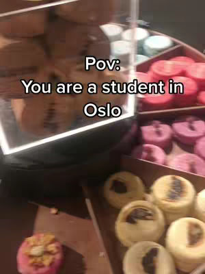 The image captures a close-up view inside a pastry shop, filled with various colorful desserts. The foreground features a selection of round, decorated pastries in a cardboard tray. The background is slightly blurred, showcasing more baked goods behind a glass display. The layout includes a mix of colors and textures, hinting at a treat-themed video.