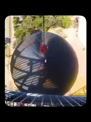 The image shows a large, dark sphere suspended from a string. A smaller, red sphere hangs below it. The background features green trees and a view from above, possibly a park environment. There is a number "3" overlaid on the sphere. The overall composition is framed with a black border.