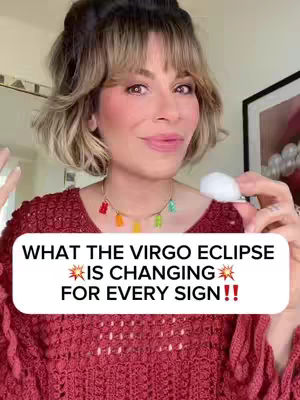 A woman with blonde hair and red sweater top is holding a white ball in the right hand. The video has a text overlay that says "WHAT THE VIRGO ECLIPSE IS CHANGING FOR EVERY SIGN!!". She is looking at the camera, smiling and speaking. She is wearing a necklace with colorful gummy bear charms.