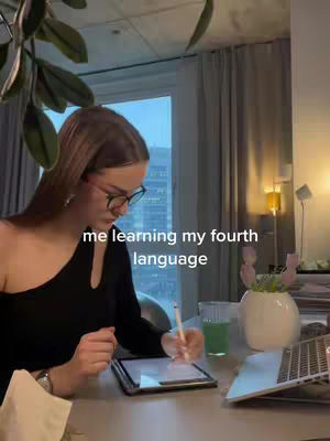 A woman with glasses is seated at a table near a window, working on a tablet. Text overlays read 'me learning my fourth language'. The background includes a window, curtains, and a few objects on the table, including a laptop and a glass of green juice.