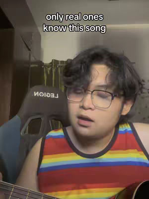 A young man with glasses is playing a guitar and singing into the camera. The background includes a black gaming chair and a curtain. The overlay text says, "only real ones know this song."