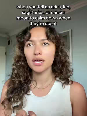 A medium shot presents a young woman with long, curly brown hair. She's positioned centrally, facing the camera with a slight upward gaze and her mouth slightly open as if speaking. She is wearing a white tank top and a delicate gold necklace. The background is plain, suggesting a home setting. The image is well-lit, with a focus on the woman's face and expression.