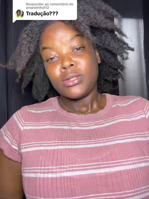 A woman with dark skin and curly black hair looks at the camera with a neutral expression. She wears a pink and white horizontally striped t-shirt and a gold necklace. The background is dark, and there is a comment bubble in the top-left corner.