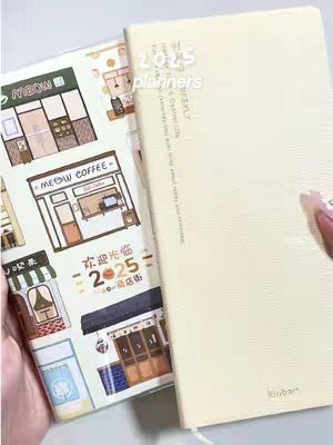 The cover image features two planners side-by-side. The planner on the left has a colorful cover with various illustrations, including a ‘MEOW COFFEE’ shop and other store-fronts and signage. The right planner is a solid, light yellow-beige color with text running down the side. A hand with manicured nails is partially visible on the right side of the image, holding the planner.