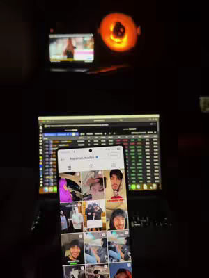 The image shows a phone displaying an Instagram feed in the foreground. Behind the phone is a laptop with a trading chart on the screen. In the background, a small television screen is visible, along with a glowing orange light, possibly from a lamp or other light source. The overall composition is dark, with the focus on the phone and laptop screens.