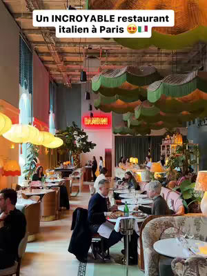 An interior shot of a restaurant. The space is filled with tables, chairs, and people dining. The lighting is warm. There are decorative ceiling elements that look like green and white clouds. A neon sign that says "Bambini" is visible. The image has an inviting, lively atmosphere.
