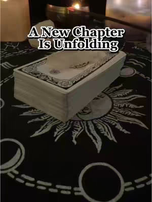 The cover image is a dimly lit scene, likely indoors, featuring a black cloth patterned with white celestial and geometric designs. On top of the cloth is a deck of tarot cards, centered and slightly angled. Above the cards, the text, 'A New Chapter Is Unfolding' dominates the top portion of the image. The background is blurred, suggesting a candle or some other light source, creating a mystical ambiance. The overall tone is evocative of spiritual or fortune-telling themes, with the high contrast between the black and off-white materials.