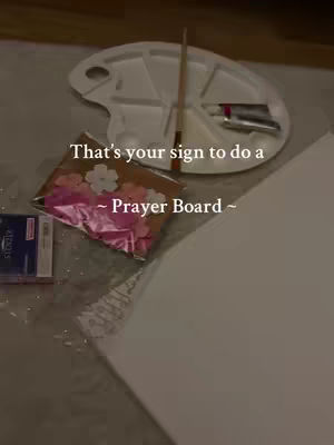 The image shows a collection of items laid out on a surface, possibly for crafting. There is a white artist's palette with paint tubes and a brush, a package of pink and white flower-shaped decorations, and a blank canvas. Text overlays read, 'That's your sign to do a Prayer Board.' The composition is lit with soft, neutral lighting, suggesting an inviting setting.