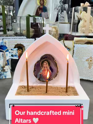 The image showcases a small, white altar with a cross-shaped top. Inside, a picture of a woman with candles is centered, surrounded by sand. Three candles are lit, with additional religious items in the background. A bright red banner at the bottom reads, "Our handcrafted mini Altars 🤍".