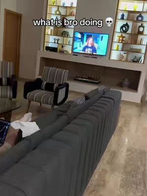 The cover image is a shot of a luxurious living room interior. The room features large, built-in shelves with decorative items and a flat-screen TV. A large gray sofa is in the foreground. The creator is partially visible, lying on the couch, holding a phone. There is additional seating with a striped chair. The color palette mainly consists of neutral tones with wooden accents, giving a sense of comfort.