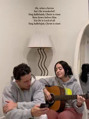 A couple is seated in a domestic setting, with the woman playing an acoustic guitar and singing. Text overlays the image containing lyrics. The scene takes place against a neutral background with a white lampshade and a curved mirror. The color palette is soft and warm, highlighting the couple and the guitar.