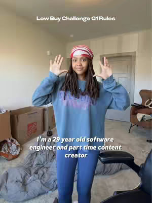 The cover image features a young woman, standing indoors with arms raised and fingers spread in a gesture of excitement or emphasis. She is wearing a scarf or headband and a blue sweatshirt and blue leggings. The background includes a messy area with boxes and a chair, in a home interior setting. Text overlays the image, providing context about a 'Low Buy Challenge' and additional personal details.
