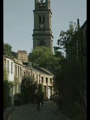An outdoor image shows a narrow street with buildings on both sides, leading towards a tall clock tower. The street is paved, and a few people are walking along it. The lighting suggests a sunny day, and the overall scene conveys a sense of history and possibly a quaint, old-world setting.