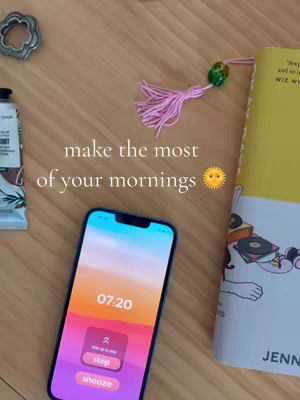 Image shows a top-down view of objects on a wooden surface. A smartphone displays an alarm screen at 7:20 am with stop and snooze buttons. Beside the phone is a partially visible book with a decorative tassel bookmark and a closed tube of cosmetic product. Text overlay in the center says make the most of your mornings with a sun emoji.