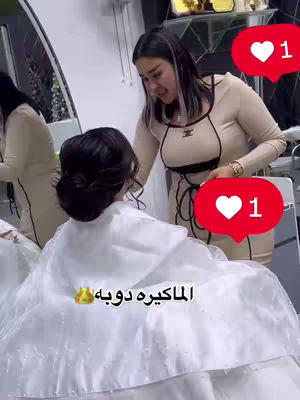 A woman in a beige dress is assisting a person who is wearing a white cloak or cape. Two red heart-shaped icons with a '1' inside are overlaid in the upper-right and lower-right corners of the image. The person being assisted has their hair up.