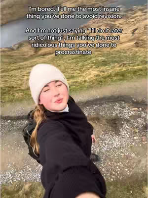 A woman in a white beanie and black jacket, with a backpack, looking at the camera. The background shows a blurred view of a natural landscape with a body of water.