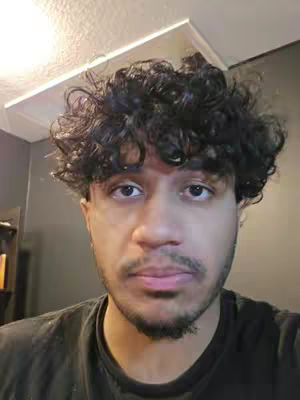 A close-up shot of a man with dark curly hair and a trimmed beard, looking directly at the camera with a neutral expression. The lighting appears to be soft and diffused from an unseen source above, casting a faint shadow on the wall behind. The background is a solid color, and the man is wearing a black t-shirt.