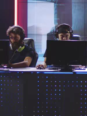 Two individuals are shown at a desk in what appears to be a gaming setup. Both are wearing headsets and focused on their computer screens. The background has blurred graphics with colors in red and blue lights.