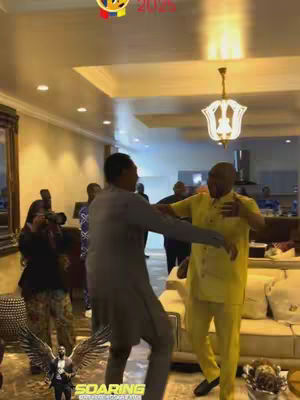 Two men embrace indoors in a living room setting, likely greeting each other. The image focuses slightly on the men, people in the background and a background with interior design elements. The setting is a living room with warm lighting. The composition is busy, and the use of a low angle shot gives a sense of intimacy.