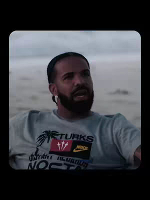 Drake is sitting on the beach in a light-colored t-shirt with text on it. The ocean and shore are in the background and the sky is overcast. The image is framed with a black border.