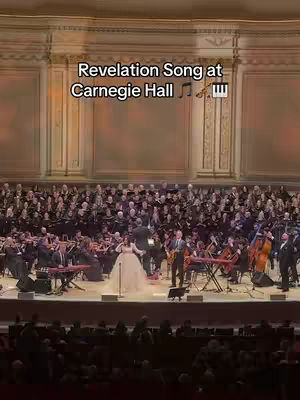 A live performance at Carnegie Hall. A woman in a white gown sings in the center stage, flanked by two men and a full orchestra and choir. The audience is visible in the foreground.