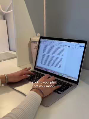 The image shows a person's hands typing on a laptop keyboard on a white desk. The laptop screen displays a document. The text overlay reads 'stick to your plan. not your mood.' A pen and a water bottle are on the right side of the laptop. The setting appears to be in a well-lit study space.